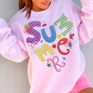 May include: A light pink sweatshirt with the word "SUMMER" in colorful, patchwork-style letters. The design includes a sun, cherries, a butterfly, and a strawberry. The sweatshirt is paired with pink shorts.
