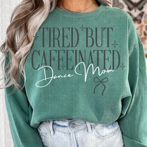 May include: A green sweatshirt with the text "TIRED BUT CAFFEINATED Dance Mom" in a mix of fonts. The sweatshirt is a solid green color and is paired with light-wash denim jeans. The text includes a bow graphic.