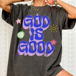 May include: Dark gray t-shirt with the words "GOD IS GOOD" in blue bubble letters outlined in purple. The shirt features a star, a smiley face, a flower, and a sun graphic.