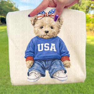 Könnte beinhalten: Eine beigefarbene Tragetasche mit einem Teddybären, der einen blauen Pullover mit dem weißen Schriftzug "USA", Jeans und weiße Turnschuhe trägt. Der Bär hat eine Schleife mit einem rot-weiß-blauen Muster. Die Tasche wird von einer Hand gehalten.