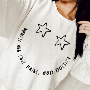 May include: White sweatshirt with a black smiley face design made of two stars and the text "WHEN ALL ELSE FAILS, GOD DOESN'T". The sweatshirt is a crew neck style.