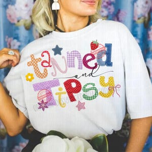 May include: White t-shirt with the words "Tanned and Tipsy" in colorful, patchwork-style letters. The design includes a strawberry, stars, and a flower. The shirt has a relaxed fit.