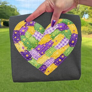May include: A dark gray tote bag featuring a large, heart-shaped appliqué. The appliqué is a patchwork design in yellow, green, and purple fabrics, with star and gingham patterns. The bag is held by a hand.