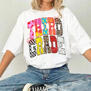 Op de afbeelding: Een wit t-shirt met korte mouwen en de tekst THIRD GRADE in grote blokletters. Elke letter heeft een ander patroon, waaronder kersen, gele vlammen, blauwe stippen, roze tijgerstrepen, luipaardprint, zwart-wit geblokt, gele sterren, camouflage en een rode dierenprint.