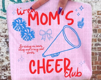 May include: A pink rectangular item with the words "tired MOM'S CHEER club" in red, and blue pom-poms, a bow, and a megaphone. The text "Surviving on bows, bling and crazy cool stunts" is also present.