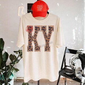 Peut inclure: Un t-shirt crème avec les lettres "KY" dans un motif léopard. Au-dessus du t-shirt, une casquette de baseball rouge avec le texte "MAD LOVE FOR YOU". Le t-shirt est suspendu à un cintre noir.