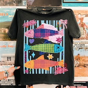 以下が含まれることがあります： カラフルな魚のデザインが施された黒いTシャツ。ピンク、青、黄色、オレンジのパッチワーク模様の魚が3匹、青と白のストライプの背景に配置されています。星、ハート、グリッターなどの装飾要素が含まれています。