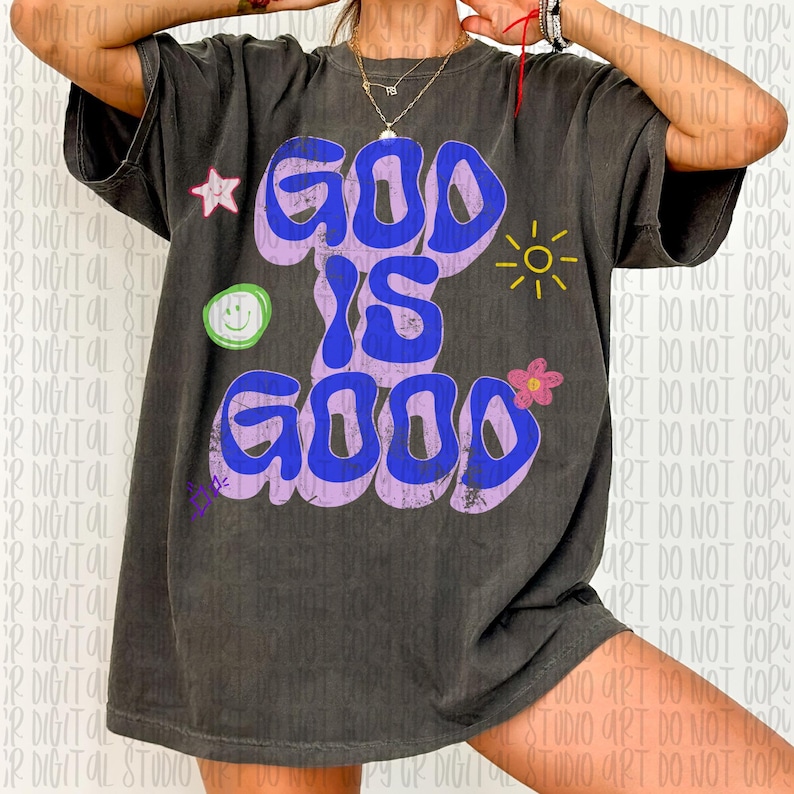 May include: Dark gray t-shirt with the words "GOD IS GOOD" in blue bubble letters outlined in purple. The shirt features a star, a smiley face, a flower, and a sun graphic.
