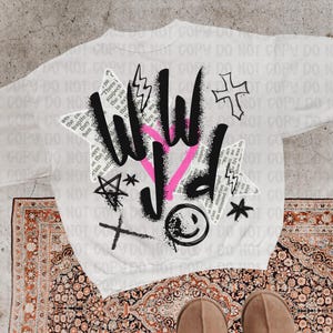 Puede incluir: Sudadera blanca con un dise&ntilde;o gr&aacute;fico que presenta las letras "WILD" en negro y rosa, junto con una cruz, rayos, estrellas y una carita sonriente. El dise&ntilde;o incorpora elementos de texto.
