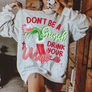 May include: A light gray sweatshirt with the text "DON'T BE A Grinch DRINK YOUR Wine". The design includes a wine glass, Grinch-themed elements, and a candy cane. The text and graphics are in red, green, and pink.