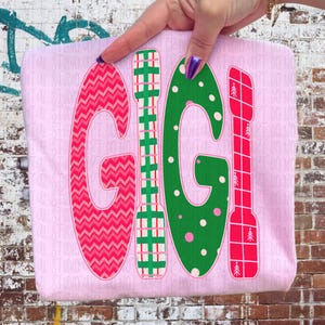 Könnte beinhalten: Ein hellrosa T-Shirt mit dem Wort "GIGI" in großen, farbenfrohen Buchstaben. Die Buchstaben sind mit verschiedenen Mustern verziert, darunter ein Zickzack-Design, grüne Punkte und rotes Karomuster. Das Design wird von einer Person gehalten.