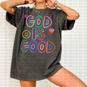 May include: Dark gray t-shirt with the words "GOD IS GOOD" in colorful, playful lettering. The design includes a smiley face, a heart, and a star. The shirt has a relaxed fit.