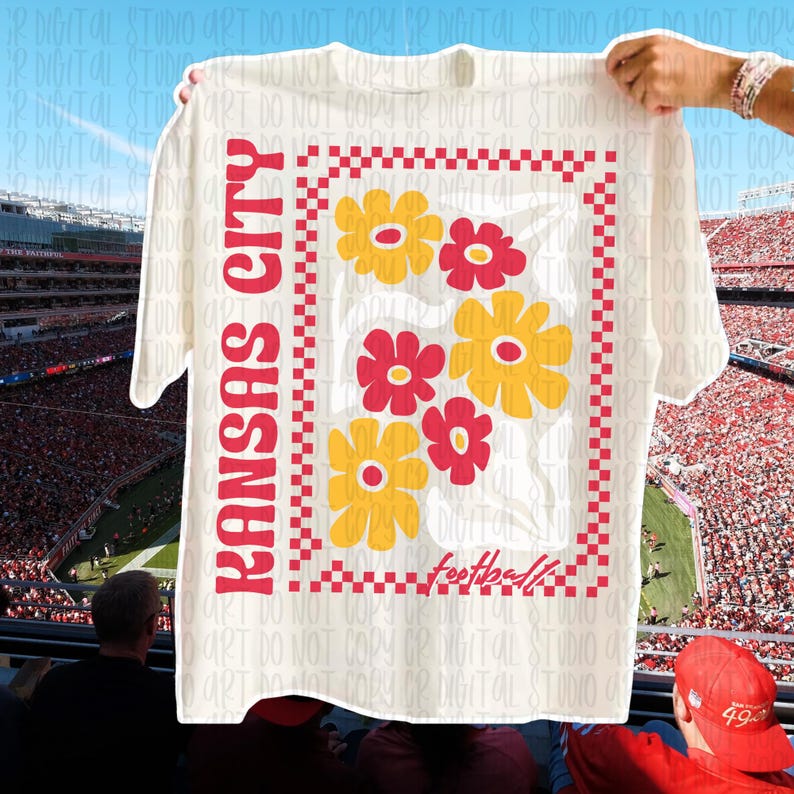 May include: Cream-colored t-shirt featuring the text "Kansas City" in red, with a floral design in red and yellow. The word "football" is written in script at the bottom of the design. The shirt is held up in front of a stadium.
