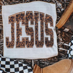 May include: A cream-coloured t-shirt with the word "JESUS" printed in large, bold letters. The letters have a leopard print pattern with a brown outline. The shirt is surrounded by other items, including a boot and a checkered pattern.