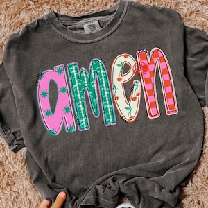 May include: Dark gray Comfort Colors t-shirt with the word "AMEN" in colorful, patterned letters. The "A" is pink with green starbursts, the "M" is green with a bamboo design, the "E" is cream with cherries, and the "N" is orange and pink checkered.
