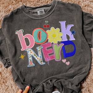 May include: Dark grey t-shirt with the words "BOOK NERD" in colorful, embroidered letters. The design includes a flower, cherries, a star, and hearts. The shirt is made of a soft material.