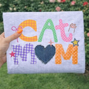 May include: A light gray t-shirt with the words "CAT MOM" in colorful, patchwork-style letters. The "C" and "A" are yellow and orange gingham, the "T" is pink gingham, and the "MOM" is purple gingham, denim, and orange polka dots. A cat paw and star accents the design.