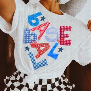 Może przedstawiać: Biała koszulka z napisem "BASEBALL" naszytym w różnych wzorach i kolorach, w tym niebieskie kropki, czerwona kratka i niebieska kratka. Koszulka posiada również niebieskie gwiazdki.