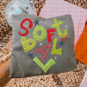 May include: A gray sweatshirt with the words "SOFTBALL" appliqued in red, green, and blue patterned fabric. The letters are outlined with white stitching and accented with small stars and plus signs.