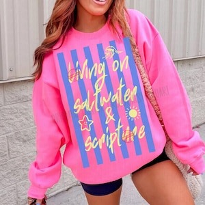 May include: A bright pink sweatshirt with the text "Living on Saltwater & Scripture" in yellow script. The design includes blue vertical stripes and small illustrations of a sun, starfish, and other elements.