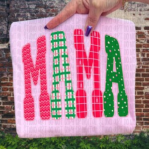 May include: A light pink t-shirt with the word "MAMA" in large, colorful letters. The letters are decorated with various patterns: red knit, green plaid, red plaid with small trees, and green polka dots. The shirt is held up by a hand.