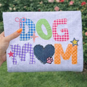 May include: A light gray t-shirt with the words "DOG MOM" appliqued in various colors and patterns. The letters are in blue gingham, green polka dots, pink gingham, purple gingham, and orange polka dots. A denim heart and paw print are also included.