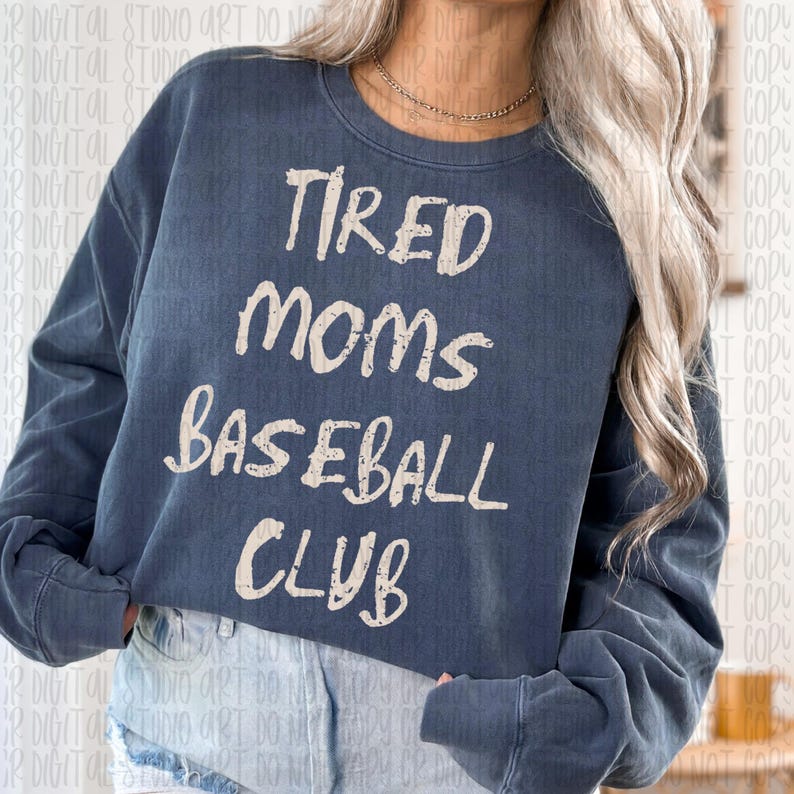 May include: A navy blue sweatshirt with the text "Tired Moms Baseball Club" in a white, handwritten font. The sweatshirt has long sleeves and a crew neck. The overall style is casual and comfortable.