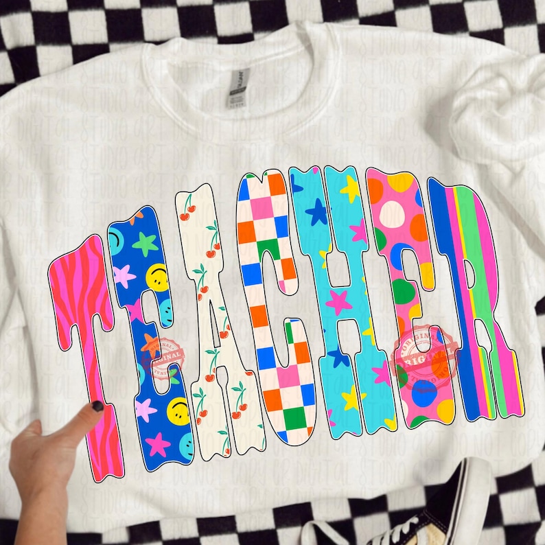 May include: White crewneck sweatshirt with the word "TEACHER" in colorful, patterned letters. The letters feature designs like zebra stripes, cherries, smiley faces, stars, and polka dots. A fun and vibrant design.