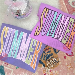 May include: Two lilac t-shirts with the word "SUMMER" in a colourful, retro font. A disco ball and a gold-coloured ring are also visible. The shirts are likely part of a summer clothing collection.