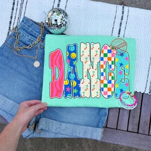 May include: A light green t-shirt with the word "DANCE" in a colorful, retro font. The letters feature patterns like smiley faces, cherries, and checkered designs. The shirt is paired with denim shorts, a gold chain necklace, and a disco ball.