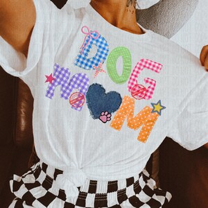 May include: White t-shirt with the words "DOG MOM" in colorful patchwork letters. The design includes a blue heart, paw print, and star accents. The shirt is knotted at the waist.