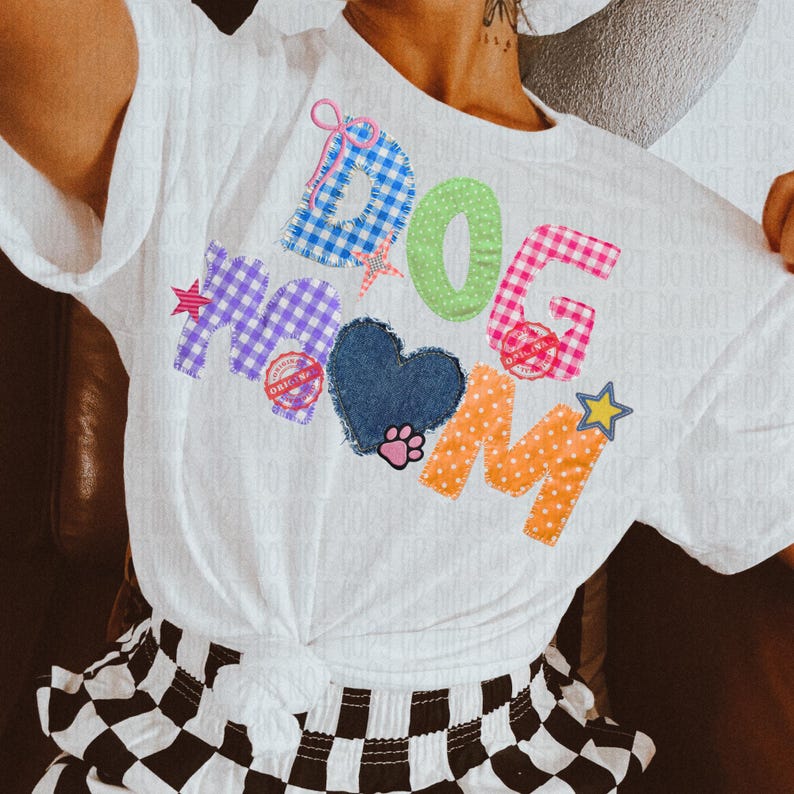 May include: White t-shirt with the words "DOG MOM" in colorful patchwork letters. The design includes a blue heart, paw print, and star accents. The shirt is knotted at the waist.