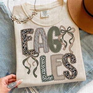 May include: Cream-colored t-shirt with the word "EAGLES" in a decorative font. The letters feature patterns like camouflage, stripes, and leopard print, with ribbon accents. A floral necklace and a brown hat are also visible.