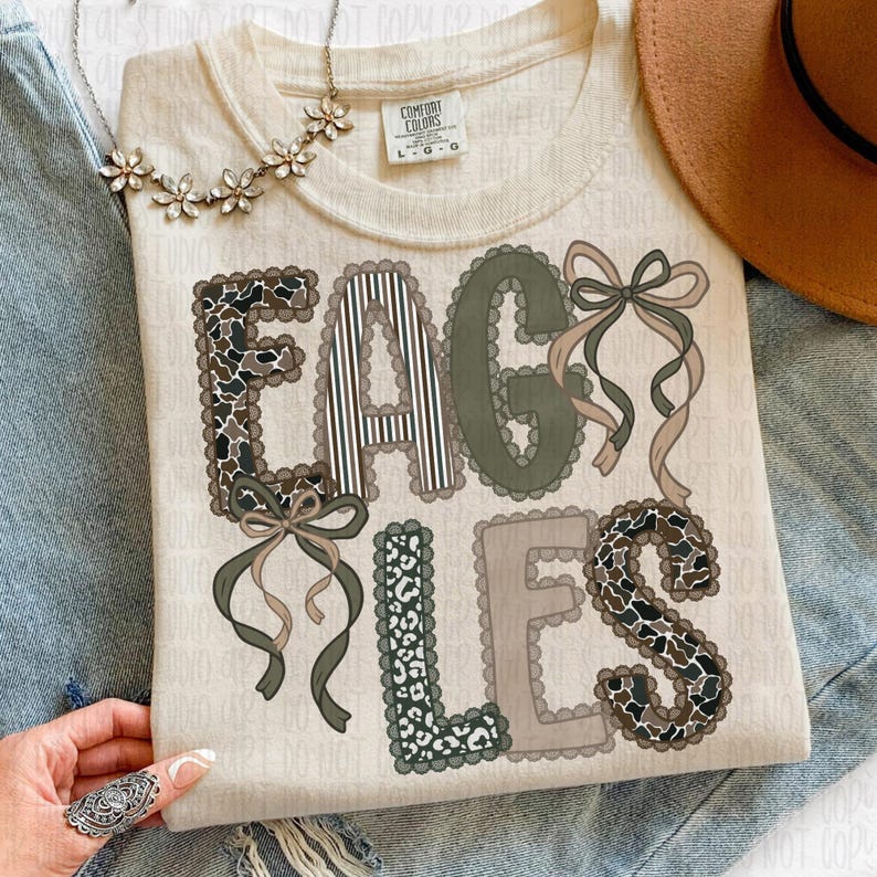 May include: Cream-colored t-shirt with the word "EAGLES" in a decorative font. The letters feature patterns like camouflage, stripes, and leopard print, with ribbon accents. A floral necklace and a brown hat are also visible.