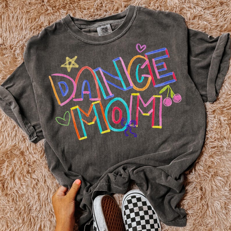 Pode incluir: Uma t-shirt cinza carv&atilde;o com as palavras "DANCE MOM" em letras coloridas desenhadas &agrave; m&atilde;o. O design inclui uma estrela, cora&ccedil;&otilde;es, cerejas e uma pequena figura de bailarina. A t-shirt &eacute; combinada com t&eacute;nis xadrez preto e branco.