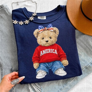 Puede incluir: Sudadera azul marino con una ilustración de un oso de peluche que lleva un suéter rojo con "AMERICA" en letras blancas, vaqueros azules y zapatillas blancas. El oso tiene un lazo con un diseño de la bandera americana. También se ve un collar plateado con detalles florales.