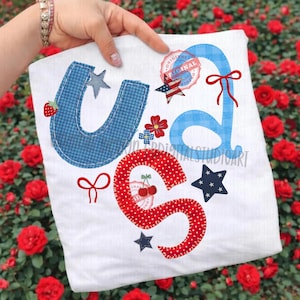 May include: A white tote bag featuring the letters "USA" in blue gingham and red polka dot fabric. The design includes stars, flowers, bows, and a strawberry and cherry illustration. The word "ORIGINAL" is printed on a circular stamp.