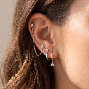 Könnte beinhalten: Ein Goldketten-Ohrring-Set bestehend aus vier Teilen. Der obere Helix-Stecker ist durch eine feine Goldkette mit einem mittleren Ohrstecker verbunden. Darunter befinden sich zwei goldene Creolen, die jeweils mit einem kleinen Drei-Stein-Anhänger verziert sind.