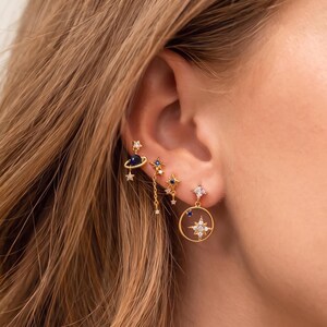 Op de afbeelding: Een set van drie goudkleurige oorbellen met hemelse motieven. Eén oorbel is een planeet met een ring, één is een hangende ketting met sterbedels en de derde is een ronde oorbel met een centrale ster en heldere kristallen.