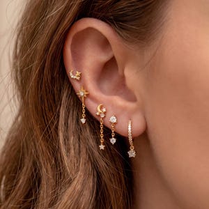 May include: A set of five gold-toned earrings featuring celestial motifs including stars, a crescent moon, and clear stones. The earrings consist of a small stud, a dangling starburst, a moon and star drop, a single stone stud, and a small hoop with a star charm.