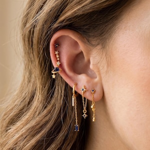 Peut inclure: Une boucle d'oreille grimpante dorée ornée d'une série de petites étoiles et d'une planète bleue, accompagnée de trois boucles pendantes. L'ensemble comprend un anneau avec une pierre baguette, un clou étoile avec chaîne et une fine chaîne étoilée.