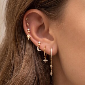 May include: A collection of gold-toned earrings featuring celestial motifs including a crescent moon, starbursts, and small clear stones. The set includes multiple stud earrings and two dangling hoop earrings arranged along the ear lobe and cartilage.