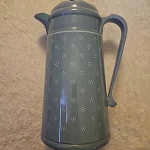 May include: A tall, light blue coffee pot with a lid and handle. The pot has a white band around the middle and is decorated with small white floral patterns. The pot is designed for serving hot beverages.