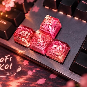 May include: Four translucent resin keycaps with a pink and red marbled pattern featuring embedded white koi fish figures. The keycaps are arranged on a black mechanical keyboard near text that reads LOFI KOI, surrounded by pink cherry blossom accents.