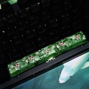 Könnte beinhalten: Ein grüner und weißer Artisan-Keycap mit einem Koi-Fisch-Design liegt auf einer schwarzen Tastatur. Der Keycap hat ein klares Harz mit grüner Basis und weißen und roten Koi-Fischen. Die Tastatur ist schwarz mit weißer Beschriftung.