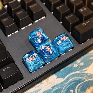 May include: Four custom resin keycaps installed on a black mechanical keyboard. Each translucent blue keycap features a detailed miniature koi fish with white and orange markings embedded within the clear, water-like textured resin material.