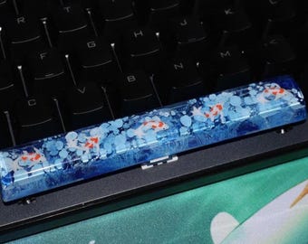 May include: A custom keyboard space bar keycap with a koi fish design. The keycap is a rectangular shape with a clear resin base, featuring a blue and white water effect with orange and white koi fish.