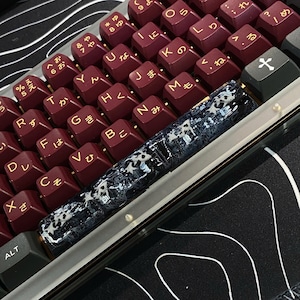 May include: A mechanical keyboard featuring deep burgundy keycaps with gold-colored Latin letters and Japanese hiragana characters. A long, translucent spacebar displays a black and white koi fish pattern embedded within the resin material.