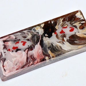 May include: Rectangular resin art piece featuring koi fish. The design incorporates white, red, and brown hues, with a glossy finish. The koi fish are depicted with red markings on a white body, set against a backdrop of swirling resin. The edges are a light gray.