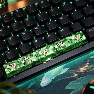 Op de afbeelding: Een spatiebalk-keycap voor een mechanisch toetsenbord met een doorschijnend groen harsontwerp met ingebedde witte en rode koi-karpermotieven. De keycap is geplaatst op een zwart toetsenbord met groene achtergrondverlichting, boven een bijpassende bureaumat.